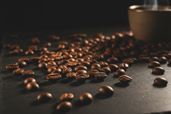 Roasted coffee beans scattered across a dark slate surface, with a blurred ceramic cup in the warm background.