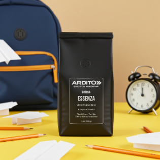A matte black coffee bag labeled ARDITO Riserva ESSENZA stands upright on a yellow tabletop, with a navy backpack, orange pencils, paper airplanes, and a black alarm clock in the background against a warm beige wall.