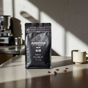 A black stand-up pouch of ARDITO Coffee Roasters labeled 'Riserva Nero' stands on a countertop in a kitchen setting, with coffee beans scattered nearby and a ceramic cup to the right. The scene is lit by strong natural sunlight with soft shadows.