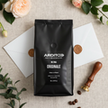 A matte black bag of ARDITO coffee beans lies centered on a light beige linen surface, surrounded by white envelopes, wax seals, coffee beans, green leaves, and peach flowers. The bag label reads 'ARDITO' and 'RISERVA ORIGINALE'.